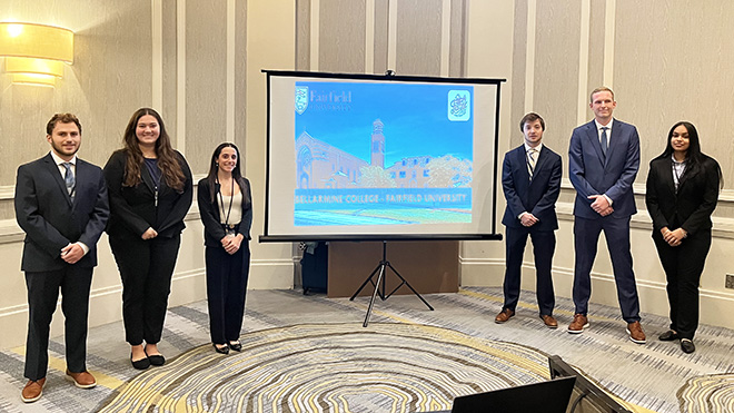 Six Roger Williams students stand next to a screen with their project slides on it