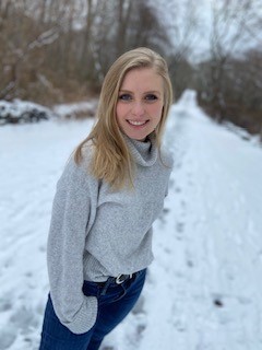 Student who identifies as female, standing in the snow, posing and smiling wearing a grey sweater with jeans. 