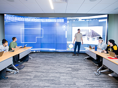 A professor stands in front of 24 television screens with a blueprint blown up on it in a classroom