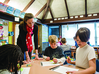 A student teacher standing next to a table with three young students 