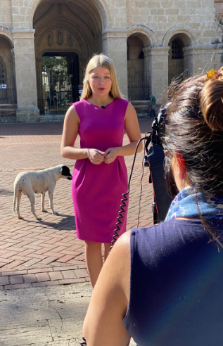 One person stands in front of building, talking to camera, while another person records. 