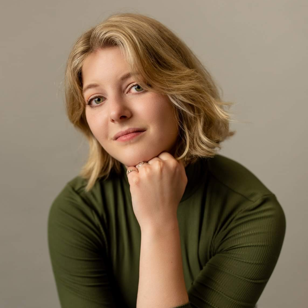A photo of Brooke Aubin. Brooke is a white, blonde person with shoulder length hair wearing a green shirt. Brooke is resting their chin on one hand and smiling slightly.