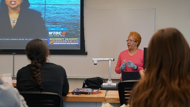 Karla-Sue Marriott stands at the front of a classroom and shows a news clip on the TV