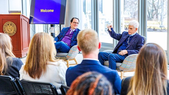 Mario J. Gabelli and Michael Melton sit in chairs in the GHH atrium in front of a crowd of students, faculty, and staff