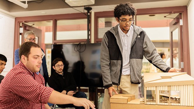 An Architecture student points at his model of a kindergarten building as a reviewer sits and gives feedback