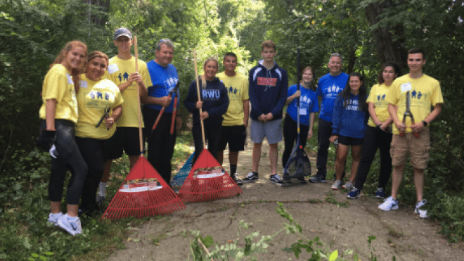 Group photo of students and president holding rakes on a path.