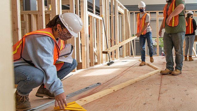 Students in hardhats, vets, safety goggles, and facemasks build a house indoors