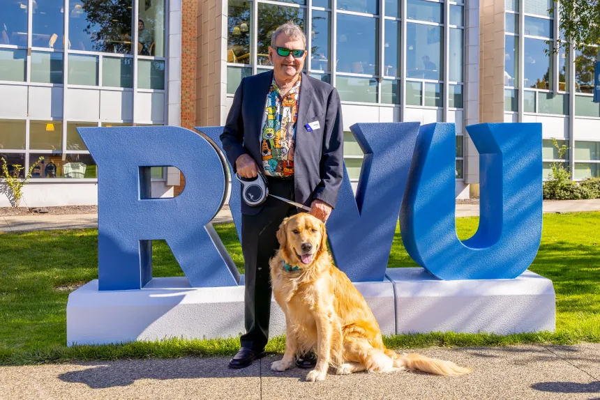 President Yannis Miaoulis and First Dog Roger pose for a photo in front of giant RWU block letters.